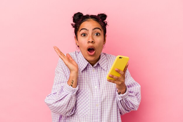 Mujer latina joven que sostiene un teléfono móvil aislado sobre fondo rosa sorprendido y conmocionado.