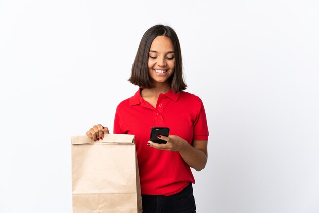 Mujer latina joven sosteniendo una bolsa de compras aislada en blanco enviando un mensaje con el móvil