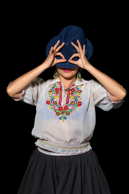 Mujer latina joven con vestido tradicional que cubre parte de su rostro con un sombrero azul en fondo negro