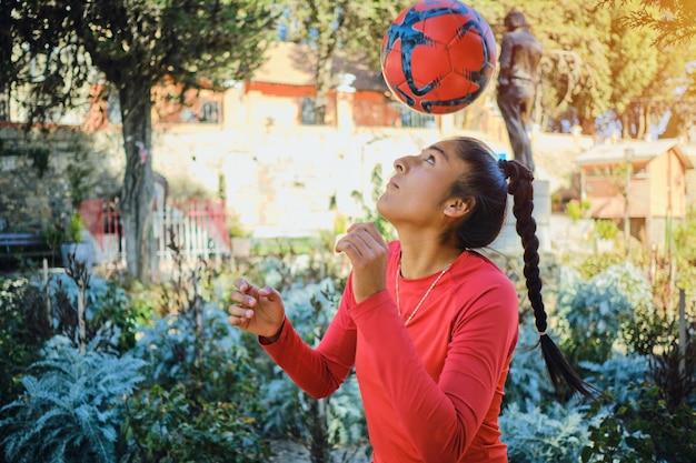 Foto mujer latina jugando al fútbol con pelota y ropa roja en un parque en bolivia américa latina