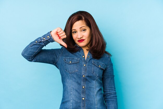 Mujer latina de mediana edad aislada en azul mostrando un gesto de aversión, pulgares hacia abajo. Concepto de desacuerdo.