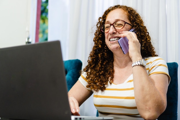 Mujer latina de mediana edad hablando por teléfono FreeLancer mientras usa su computadora en casa
