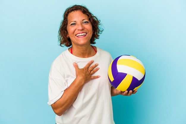 Mujer latina de mediana edad jugando voleibol aislado sobre fondo azul se ríe a carcajadas manteniendo la mano en el pecho.