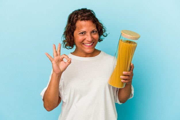 Mujer latina de mediana edad sosteniendo frasco de pasta aislado sobre fondo azul alegre y confiado mostrando gesto ok.