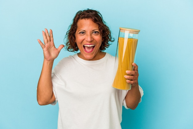 Mujer latina de mediana edad sosteniendo frasco de pasta aislado sobre fondo azul recibiendo una agradable sorpresa, emocionada y levantando las manos.