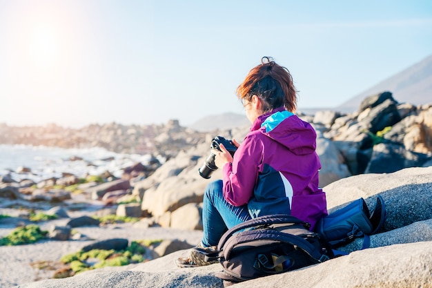 Mujer latina con mochila tomando fotografías en la playa rocosa