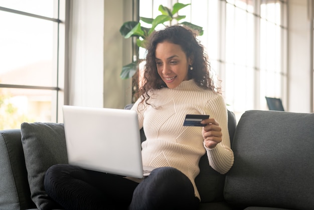 Mujer latina con ordenador portátil y mano sujetando la tarjeta de crédito para ir de compras en el sofá en casa