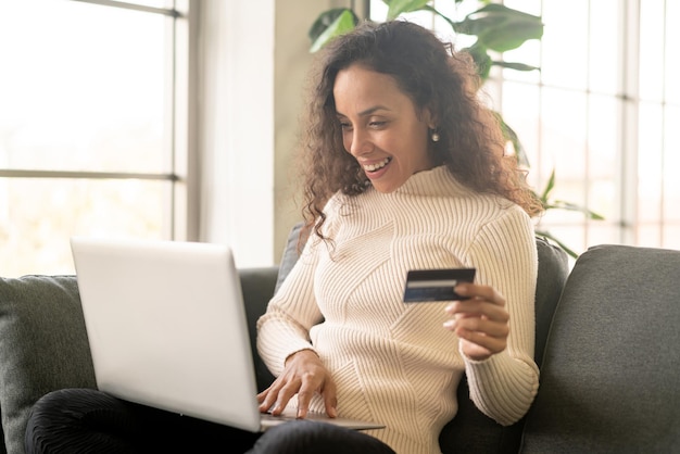 Mujer latina con ordenador portátil y mano sujetando la tarjeta de crédito para ir de compras en el sofá en casa
