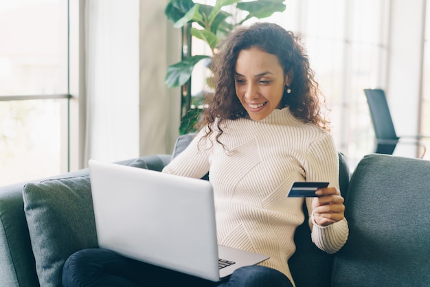 Mujer latina con ordenador portátil y mano sujetando la tarjeta de crédito para ir de compras en el sofá en casa
