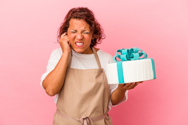 Mujer latina de pastelería de mediana edad sosteniendo un pastel aislado sobre fondo rosa cubriendo las orejas con las manos.