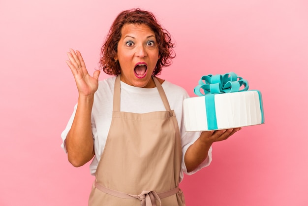 Mujer latina de pastelería de mediana edad sosteniendo un pastel aislado sobre fondo rosa sorprendido y consternado.