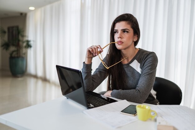 Mujer latina pensativa sentada en la mesa con el portátil pensando en la inspiración soñando