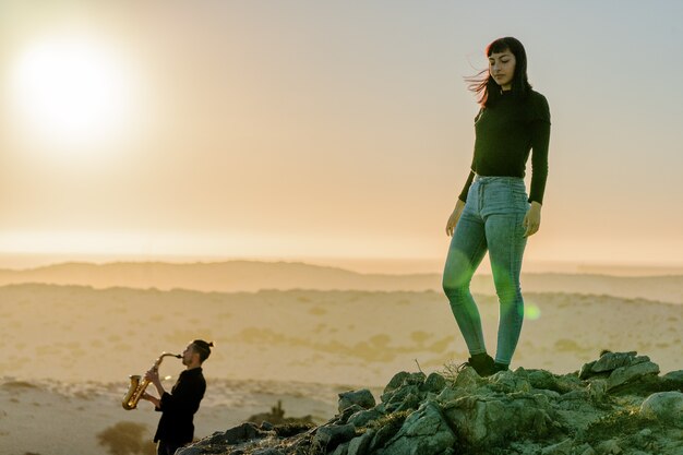 Mujer latina de pie sobre la roca mirando hacia el atardecer mientras un músico toca el saxofón