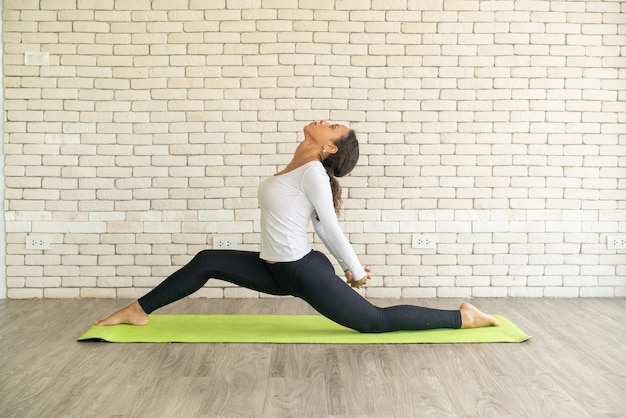 Mujer latina practicando yoga en la alfombra con fondo de ladrillo blanco