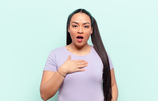 Mujer latina que se siente consternada, asombrada y sorprendida, con la mano en el pecho y la boca abierta, diciendo ¿quién, yo?