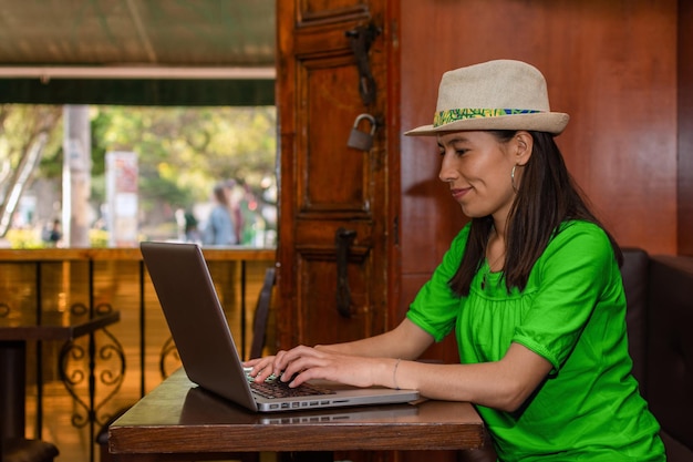 Mujer latina con sombrero y blusa verde sentada en una cafetería trabajando en su laptop