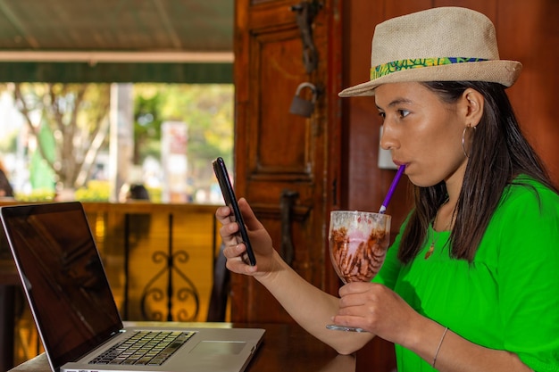 Mujer latina con sombrero y blusa verde sentada en una vieja cafetería revisando su teléfono celular mientras bebe un batido con pajita