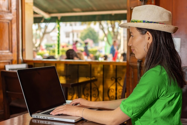 Mujer latina con sombrero y blusa verde sentada en una vieja cafetería trabajando en su laptop