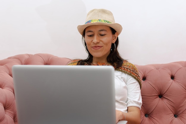 Mujer latina con sombrero sentada en un sofá rosa trabajando en su laptop