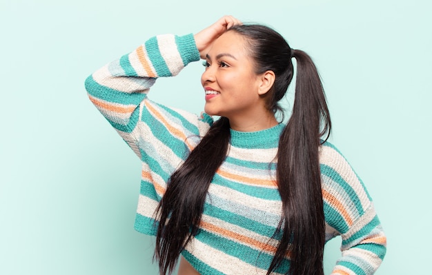 Mujer latina sonriendo alegre y casualmente, tomando la cabeza con una mirada positiva, feliz y segura