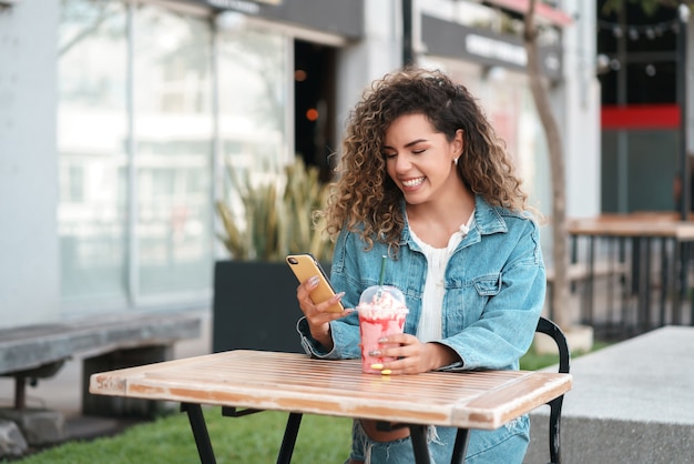 Mujer latina con su teléfono móvil mientras bebe una bebida fría en una cafetería al aire libre en la calle. Concepto urbano.