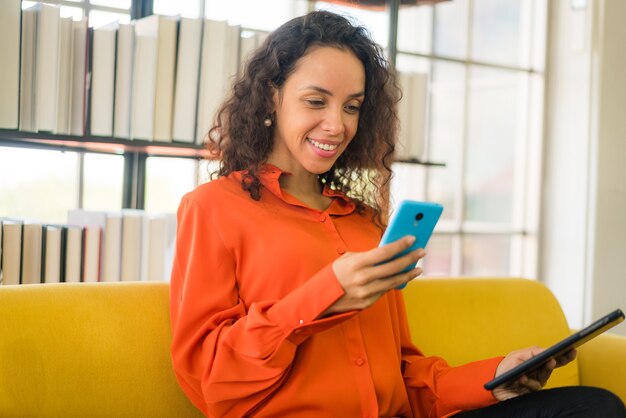 Mujer latina con teléfono móvil o smartphone y sosteniendo tablet oat home