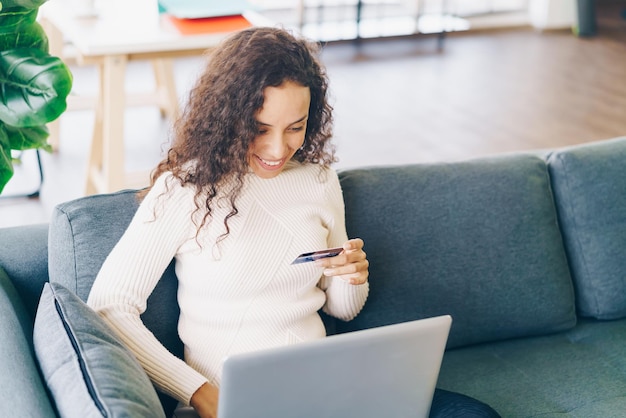 Mujer latina usando laptop y mano con tarjeta de crédito para ir de compras en el sofá