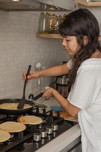 Mujer latina vestida casualmente en la cocina revolviendo huevos con queso y calentando arepas colombianas