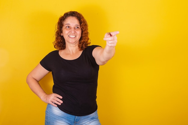 Mujer latinoamericana brasileña caucásica cabello rizado rizos fondo amarillo con el dedo apuntando a algo lejano indicando sugiriendo