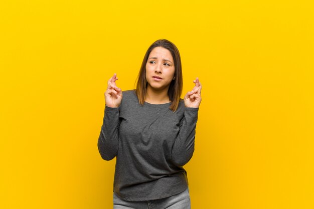 Mujer latinoamericana cruzando los dedos con ansiedad y esperando buena suerte con una mirada preocupada aislada en la pared amarilla