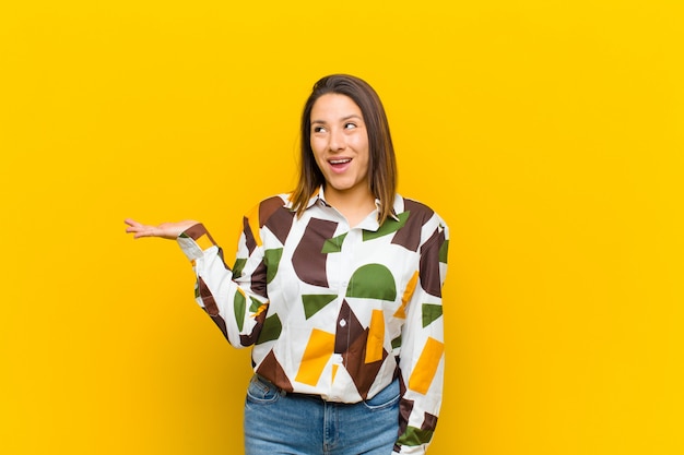 Mujer latinoamericana que parece sorprendida y conmocionada, con la mandíbula caída sosteniendo un objeto con la mano abierta en el lado aislado en la pared amarilla