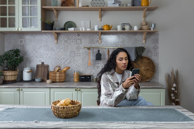 Mujer latinoamericana seria y pensativa en la cocina usa el teléfono mujer lee cuidadosamente un