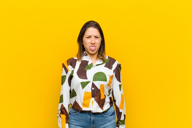 Mujer latinoamericana sintiéndose disgustada e irritada, sacando la lengua, no le gusta algo desagradable y asqueroso aislado contra la pared amarilla