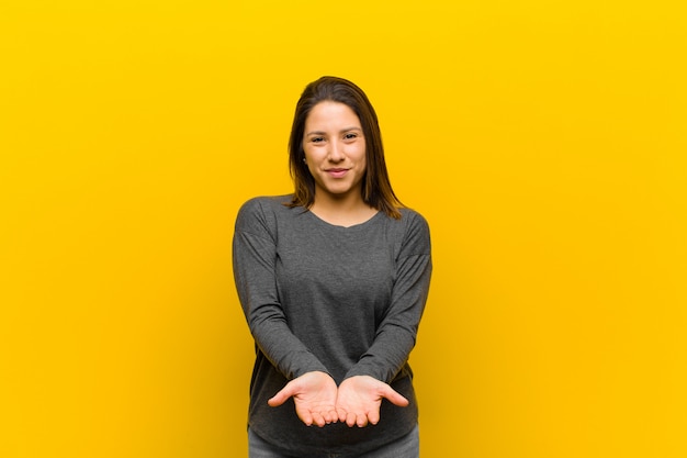 Mujer latinoamericana sonriendo felizmente con mirada amigable, segura y positiva, ofreciendo y mostrando un objeto o concepto aislado contra la pared amarilla