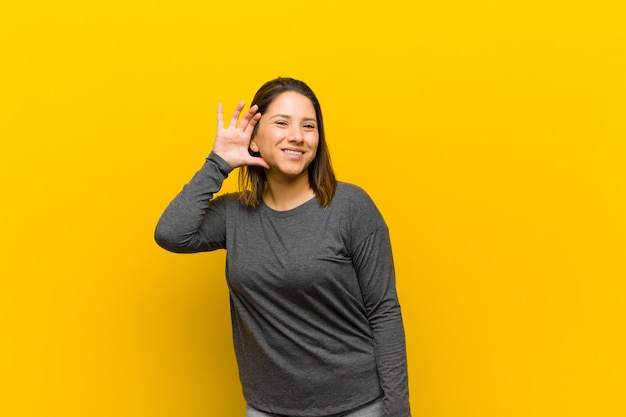 Mujer latinoamericana sonriendo, mirando curiosamente a un lado, tratando de escuchar chismes o escuchando un secreto aislado en la pared amarilla