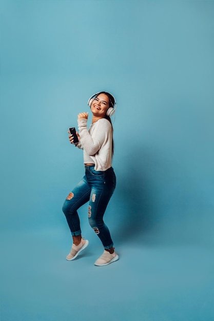 Mujer latinx bailando escuchando música con teléfono y auriculares inalámbricos sobre fondo azul.