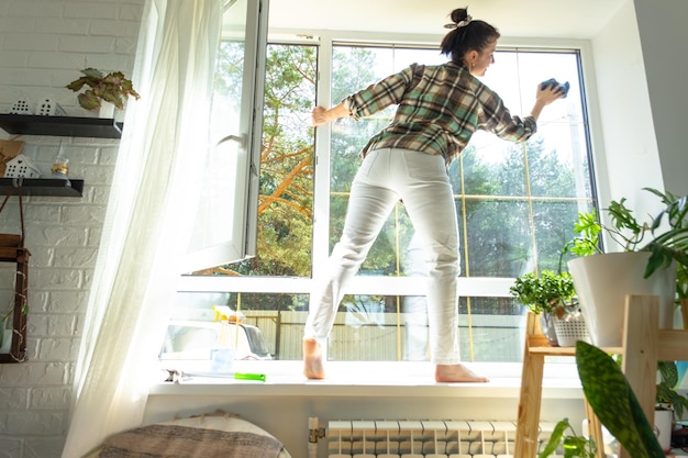 La mujer lava manualmente la ventana de la casa con un trapo con limpiador en aerosol y un trapeador dentro del interior con cortinas blancas Restaurando el orden y la limpieza en el servicio de limpieza de primavera