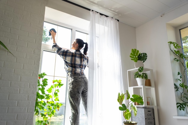 La mujer lava manualmente la ventana de la casa con un trapo con limpiador en aerosol y un trapeador dentro del interior con cortinas blancas Restaurando el orden y la limpieza en el servicio de limpieza de primavera