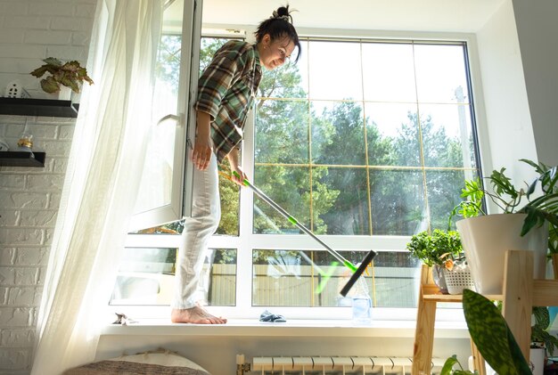 La mujer lava manualmente la ventana de la casa con un trapo con limpiador en aerosol y un trapeador dentro del interior con cortinas blancas Restaurando el orden y la limpieza en el servicio de limpieza de primavera