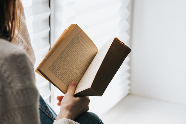 La mujer lee un libro en casa cerca de la ventana durante el día.