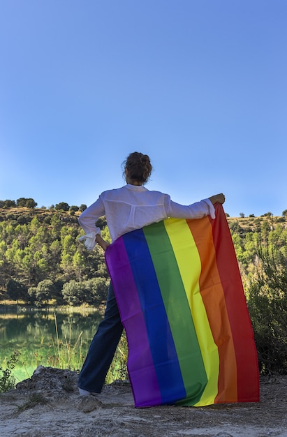 Mujer lesbiana de mediana edad irreconocible sosteniendo la bandera del arco iris gay en el lago al aire libre. Concepto de libertad