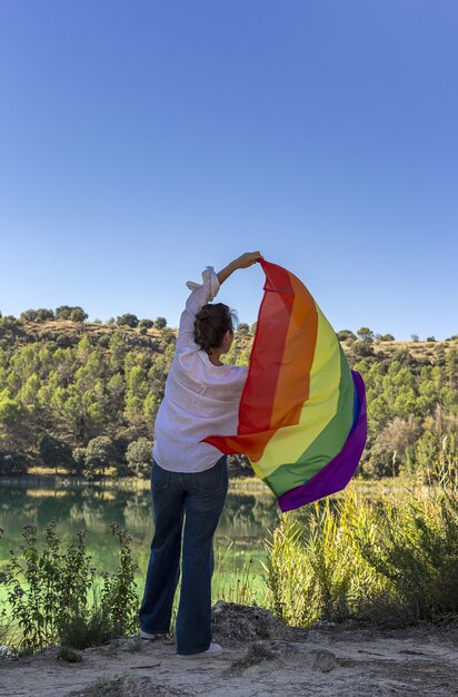 Mujer lesbiana de mediana edad irreconocible sosteniendo la bandera del arco iris gay en el lago al aire libre. Concepto de libertad