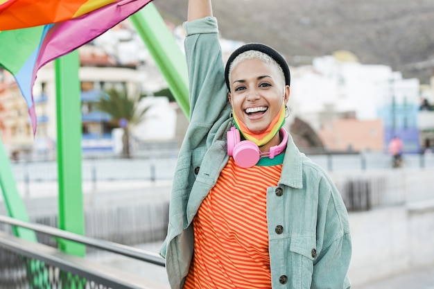 Mujer lesbiana de moda sosteniendo la bandera del arco iris lgbt en el desfile del orgullo gay mientras usa una mascarilla de seguridad debajo de la barbilla para el brote de coronavirus