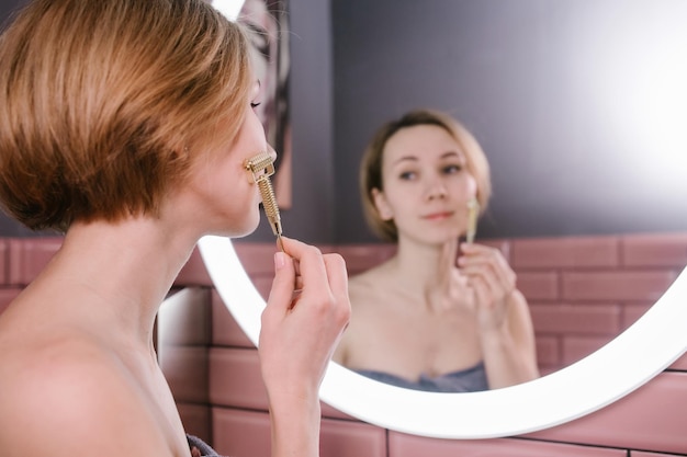 Mujer levantada masajeando su rostro con una piedra de río gua sha frente a un espejo de baño Diseño colorido de las paredes Sonriendo suavemente