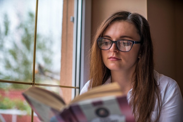 Una mujer leyendo un libro junto a una ventana.