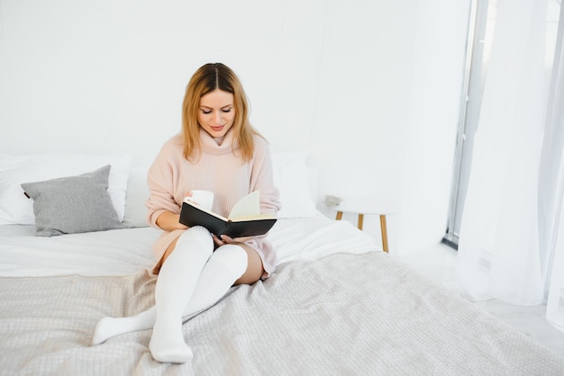 Una mujer leyendo un libro y sonriendo mientras se sienta en la cama. El despertador en el escritorio a su lado.