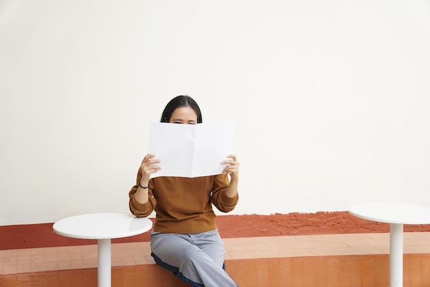 Una mujer leyendo un periódico frente a una pared blanca.