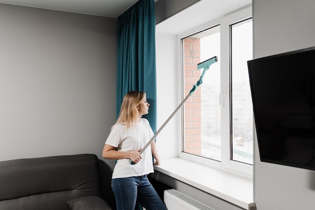 Mujer limpiadora con trapeador está limpiando ventanas en casa Servicio de limpieza de ventanas Ama de casa está haciendo tareas domésticas y lavando ventanas interiores en el apartamento