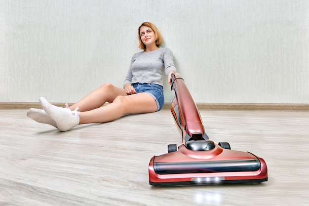 Mujer de limpieza cansada está sentada en el piso y limpiando el polvo con una moderna aspiradora vertical roja con luces LED encendidas.
