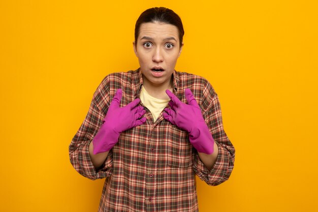 Mujer de limpieza joven en camisa a cuadros en guantes de goma mirando al frente sorprendido apuntando a sí misma de pie sobre la pared naranja